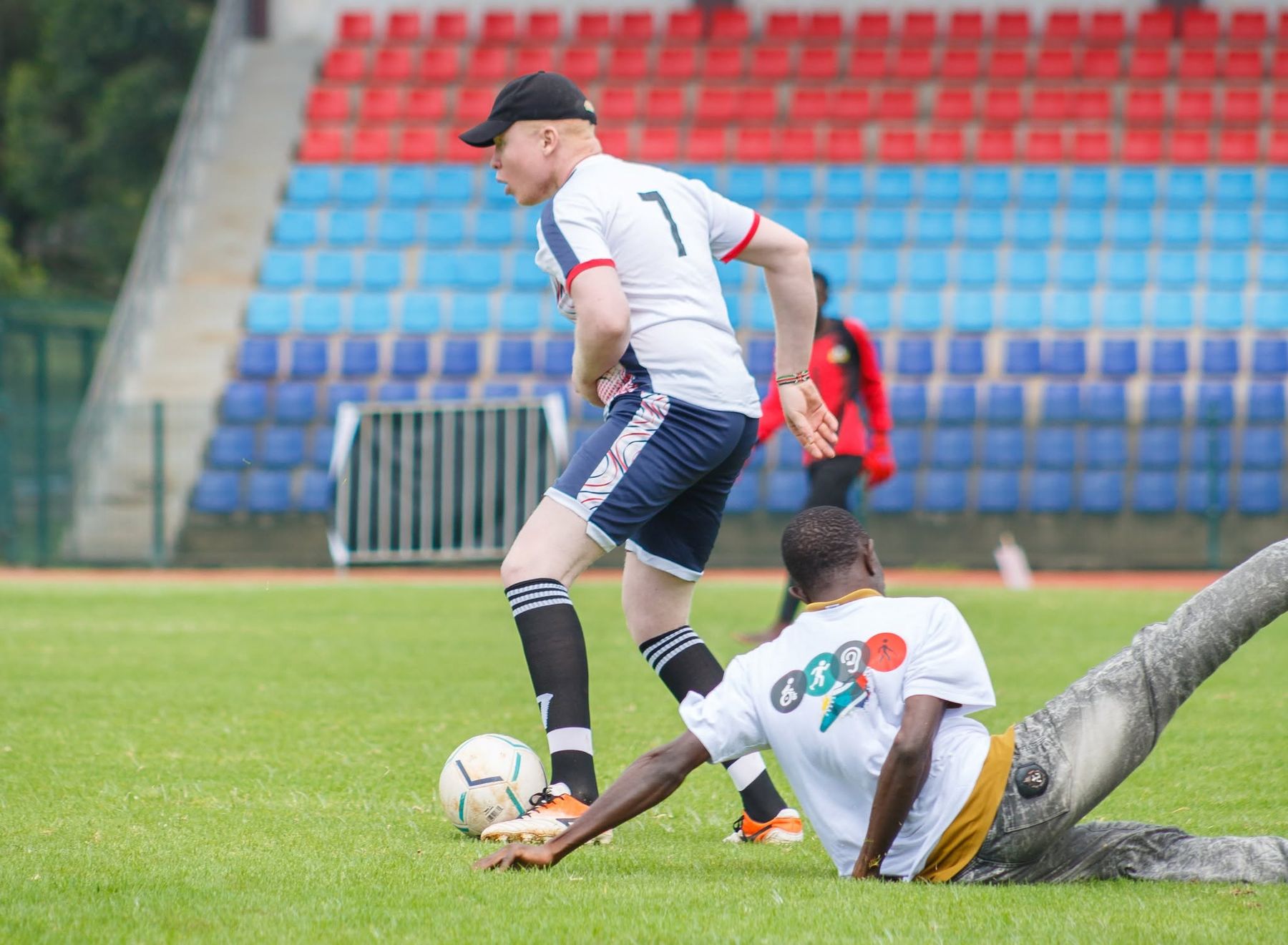 A Black Albinism footballer in a white number seven shirt and cap plays the ball past a sliding defender on a grass pitch.