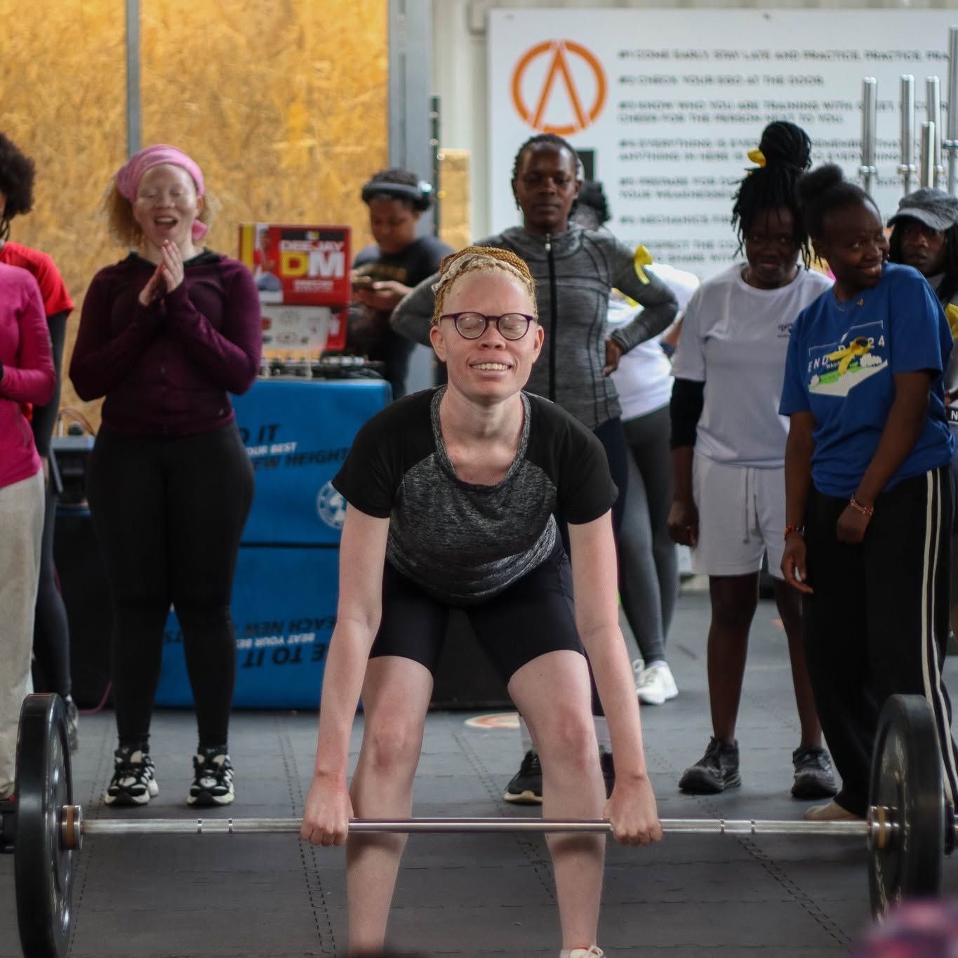 A young woman with albinism lifts a loaded barbell in a gym while other athletes watch from behind her.