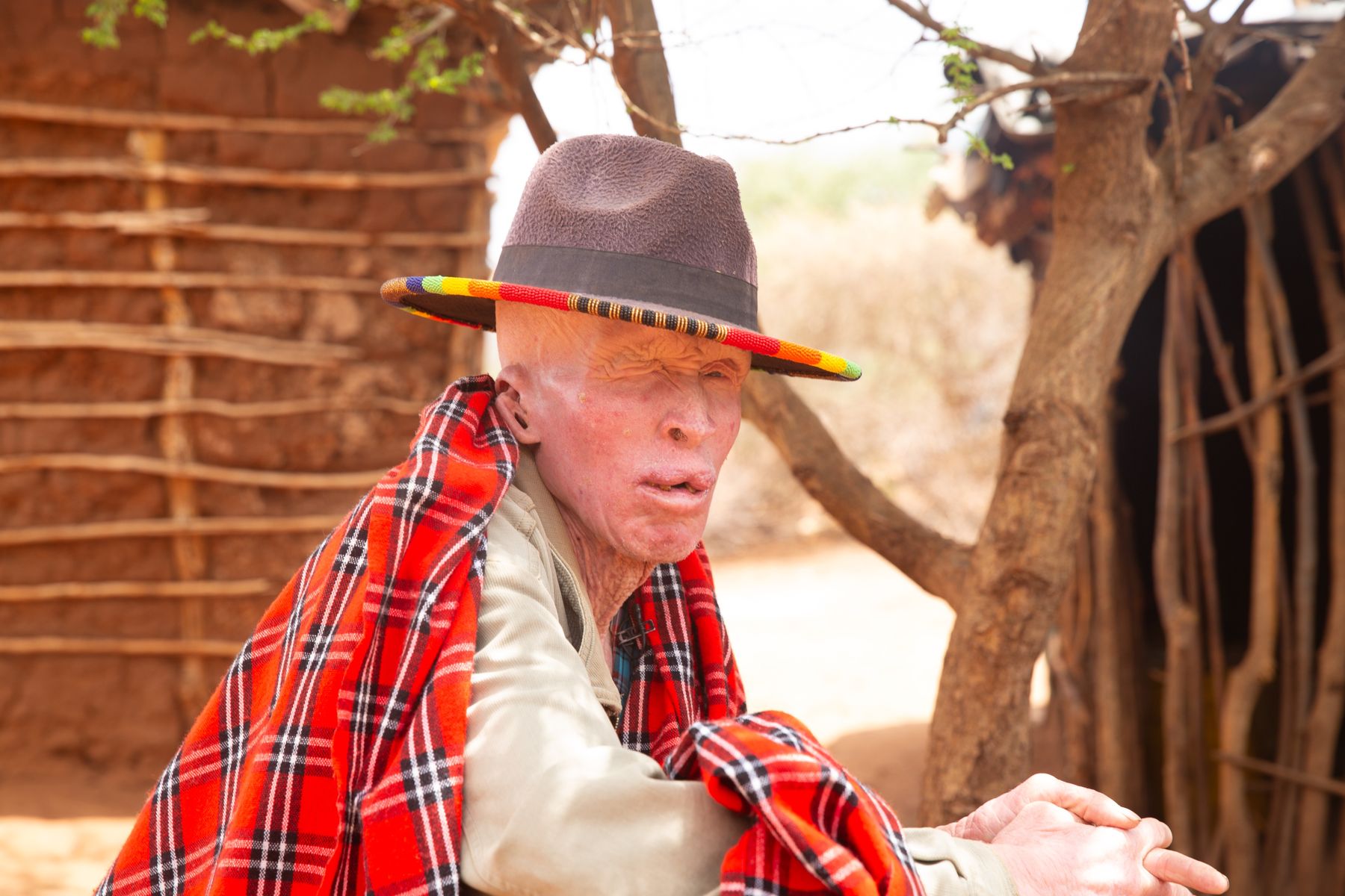 An elder man with albinism in a Maasai shuka and a wide-brimmed hat, outside his home.