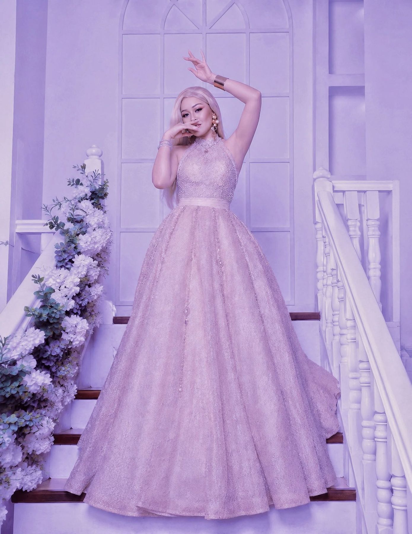A woman with albinism in a long pale gown stands on a staircase with one arm raised, mid performance.